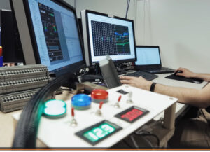 SUNCAR AG Technologiepartner für elektrische Off-Highway Systeme Engineer sits at multi-monitor workstation monitoring graphs and data on screens; foreground shows illuminated control panel with buttons and digital readouts
