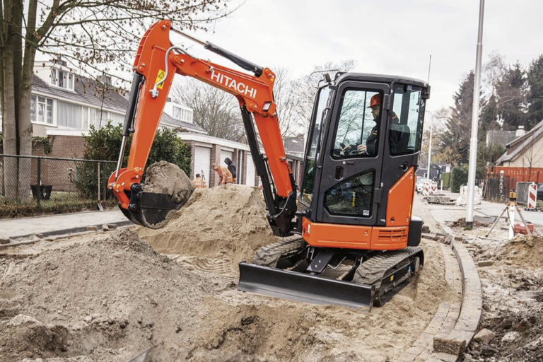 Orange Hitachi mini excavator digging a trench on a residential street with a sand pile nearby and curb work visible.