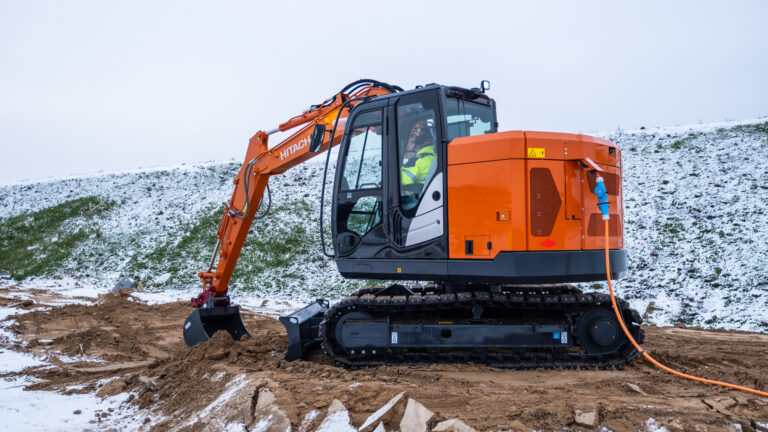 Orange Hitachi hydraulic excavator with cabin and tracked undercarriage on a mud and snow construction site, with excavator arm extended and bucket on the ground.