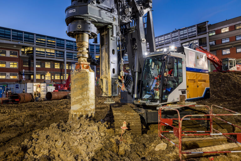 Large drilling rig on a construction site with muddy ground and a modern office building in the background