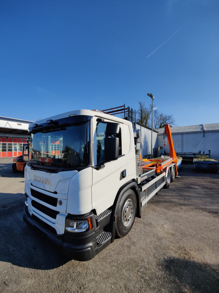 White Scania truck with a flatbed trailer carrying orange equipment in an industrial yard under a clear blue sky.