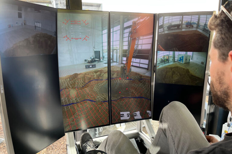 Person in a cab monitoring four screens showing a construction site with an orange excavator and a trench grid.