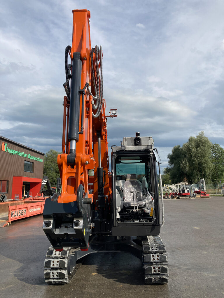 Referenzprojekt der SUNCAR AG Ihrem Technologiepartner für elektrische Off-Highway Systeme Orange hydraulic excavator arm with tracks in a construction yard, cab visible at lower right, cloudy sky overhead.