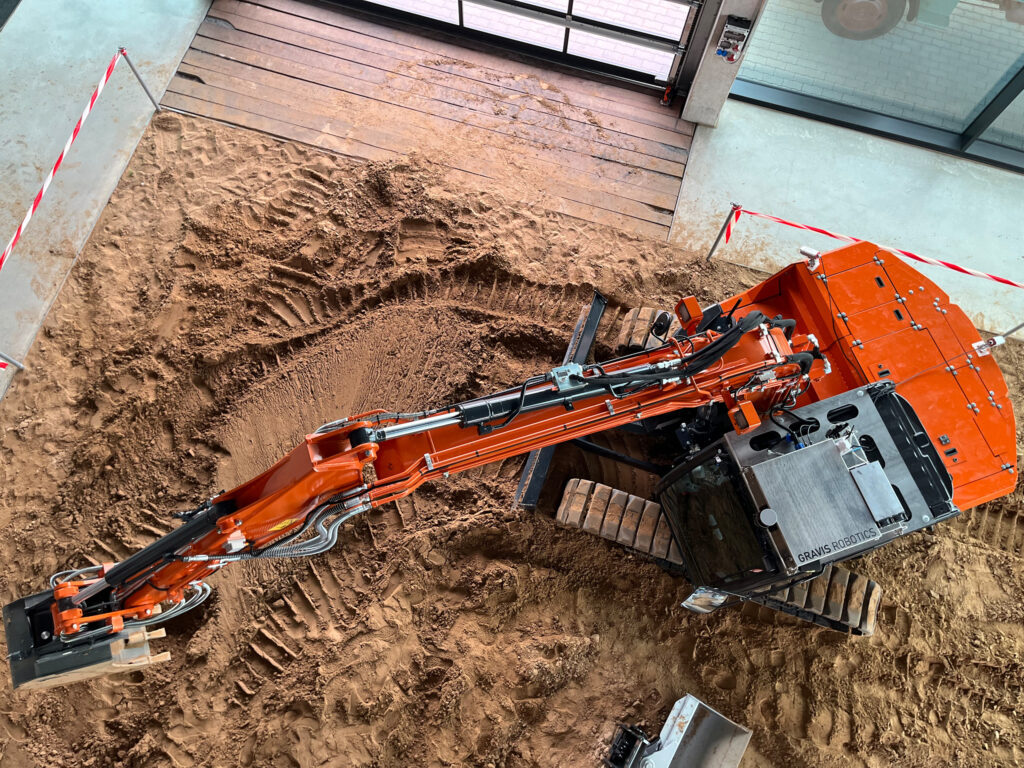 Referenzprojekt der SUNCAR AG Ihrem Technologiepartner für elektrische Off-Highway Systeme Construction excavator with orange arm digging a trench in sandy soil next to a building with large glass doors and a wooden deck nearby