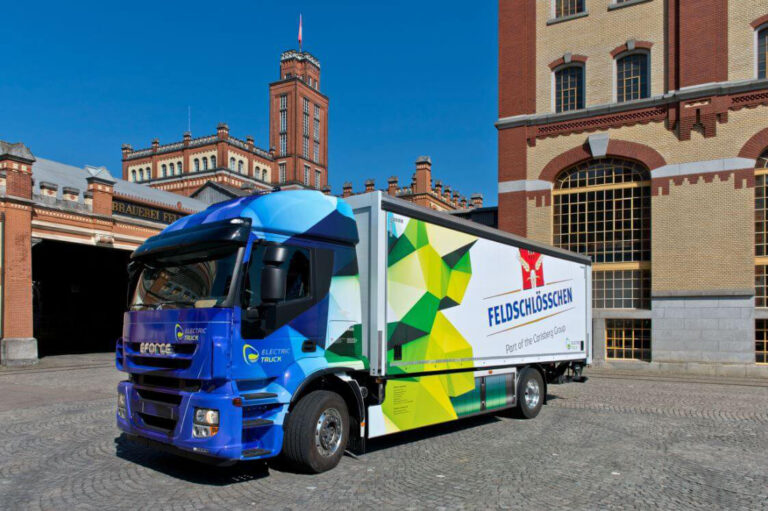 Bright blue delivery truck with a geometric green-yellow trailer parked in a historic brick square, featuring Feldschlösschen branding on the side panel.