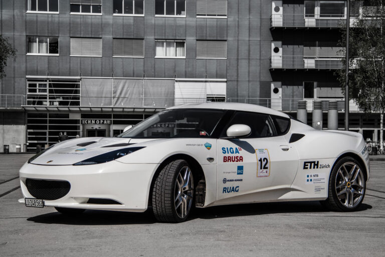 White race car with sponsor decals and number 12 parked in front of a grey modern building façade.