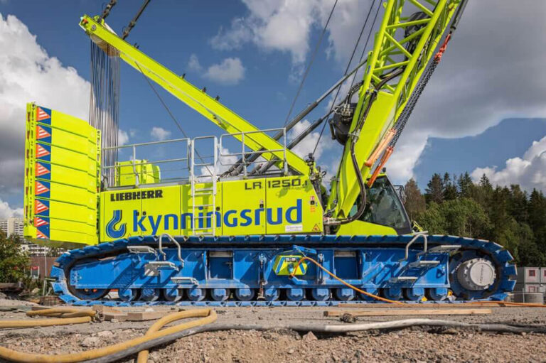 Liebherr LR 1250 crawler crane in lime-yellow with blue tracked undercarriage on a construction site, branding visible against a partly cloudy sky and trees behind.