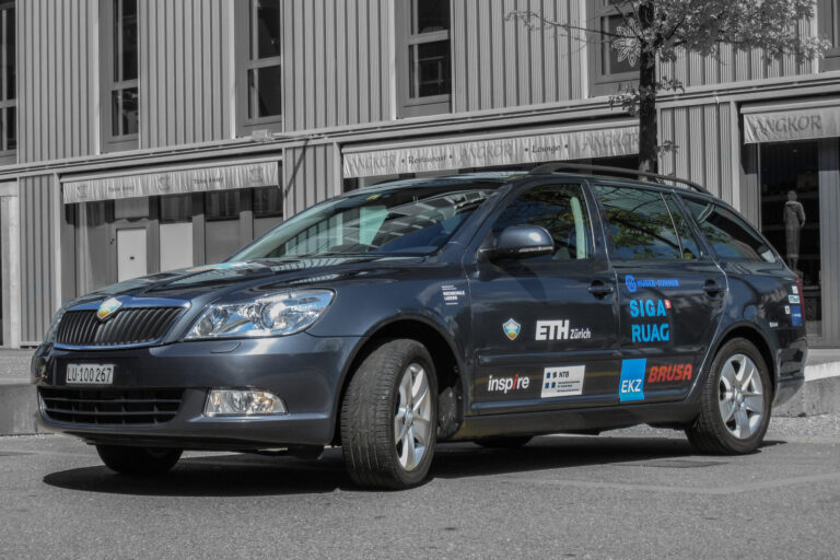 Blue SUV parked in front of a modern striped building, covered in sponsor decals including ETH Zürich and rig logos, reflecting sunlight on its hood.