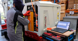 Technician in protective gear repairing a large industrial battery pack, with a laptop and toolkit on a nearby workbench.
